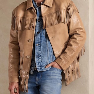 May include: Tan leather jacket with fringe detailing. The jacket has a classic collar, button closure, and pockets. Worn over a denim jacket and blue jeans, this jacket offers a stylish, Western-inspired look.