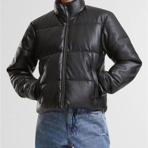 May include: A black puffer jacket with a high collar and a full-length zipper. The jacket has a quilted design and two side pockets. The model is wearing blue jeans. The jacket is made of a shiny material.