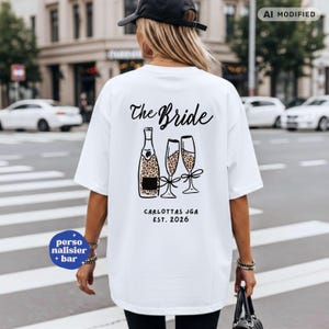 Könnte beinhalten: Ein weißes T-Shirt mit einer schwarzen Grafik auf der Rückseite, die den Schriftzug The Bride, eine Champagnerflasche und zwei Sektgläser mit Leopardenmuster zeigt. Darunter steht der Text CARLOTTAS JGA EST. 2026 in einfacher schwarzer Schrift.