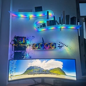 Puede incluir: Una configuración de computadora personalizada con un monitor curvo, teclado y ratón sobre un escritorio de madera. Arriba, una carcasa de PC en forma de guitarra y estantes con iluminación LED arcoíris muestran componentes y accesorios.