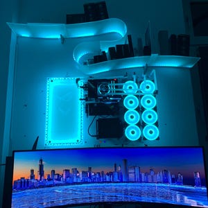 Puede incluir: Una configuración de computadora personalizada con iluminación LED azul. Los componentes están montados en una pared, incluyendo un monitor grande que muestra un paisaje urbano. La configuración presenta múltiples ventiladores y un diseño de estante único con iluminación integrada.