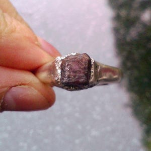 Könnte beinhalten: Ein Silberring mit einem großen, grob geschliffenen, dunkelrosa Edelstein. Der rechteckige Edelstein ist in einer einfachen Silberfassung gefasst. Der Ring wird vor einem verschwommenen Hintergrund gehalten.