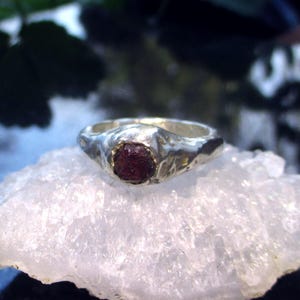 Könnte beinhalten: Ein silberner Ring mit einem strukturierten Band und einem dunkelroten Edelstein. Der Ring liegt auf einer weißen, kristallinen Oberfläche mit einem verschwommenen grün-blauen Hintergrund.