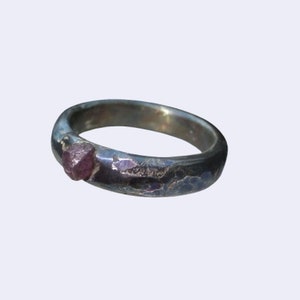 Puede incluir: Un anillo de plata con una banda texturizada y una pequeña gema de rubí en bruto. El rubí es de un color rojo rosado intenso y está engastado en la banda. El anillo tiene un aspecto rústico y artesanal.