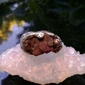 Peut inclure: Une bague avec une pierre précieuse brute rouge foncé sertie dans une bande en argent texturée à la forme organique. Le métal présente un aspect fondu et irrégulier qui entoure la pierre facettée, créant un contraste entre l'argent et le rouge.