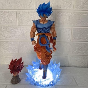 Puede incluir: Figura detallada de un personaje con pelo azul puntiagudo, vistiendo un atuendo azul y naranja, y de pie sobre una base de cristal azul brillante. También hay una figura más pequeña con pelo rojo. Las figuras están colocadas contra una pared de ladrillos blancos.