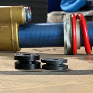 Puede incluir: Primer plano de un amortiguador de motocicleta con cuerpo azul, componentes dorados y un muelle helicoidal rojo. Dos pequeños casquillos de goma negros están en primer plano. El amortiguador está diseñado para uso de alto rendimiento.