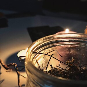 May include: A clear glass jar filled with dried moss and twigs, illuminated by the warm glow of a lit candle. The scene is set against a dark background, creating a cozy and intimate atmosphere.