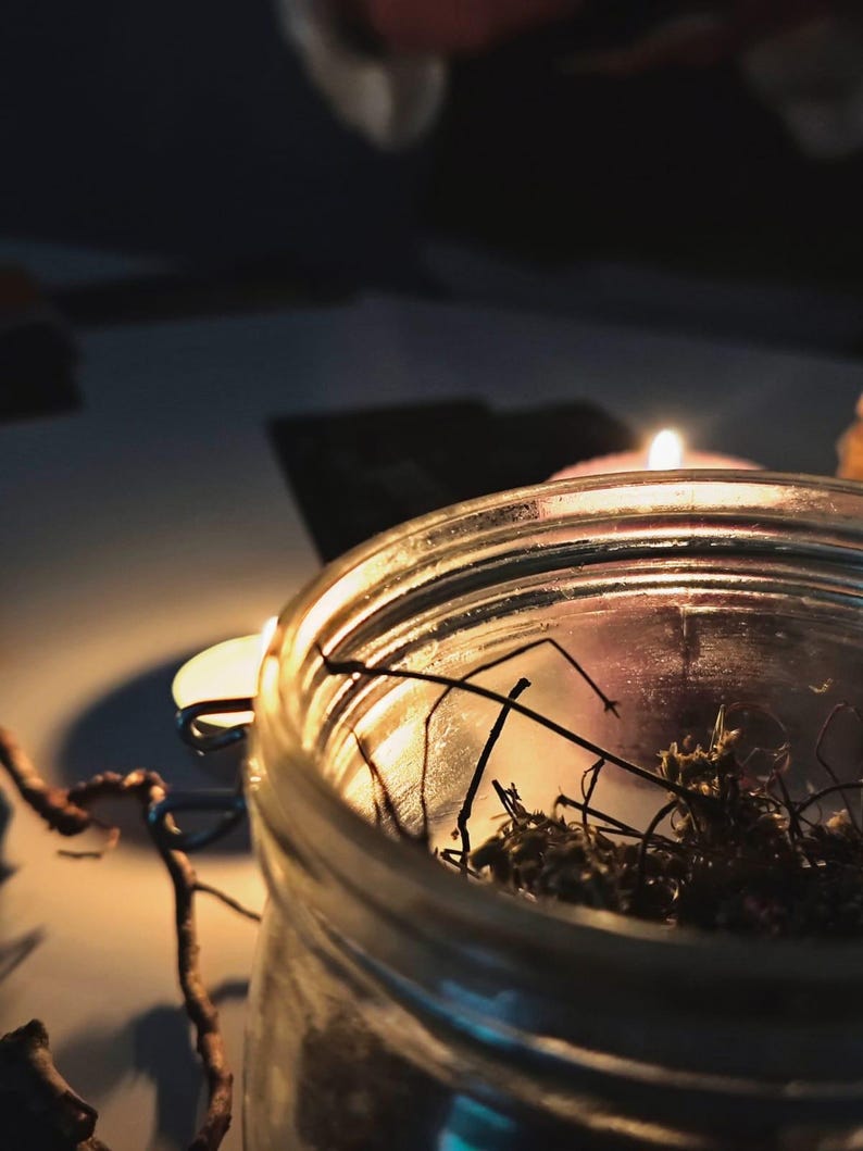May include: A clear glass jar filled with dried moss and twigs, illuminated by the warm glow of a lit candle. The scene is set against a dark background, creating a cozy and intimate atmosphere.