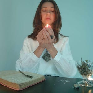May include: A woman in a white blouse holds a lit candle between her hands. An open book with a decorative bookmark and a glass bowl with dried herbs sit on a black table. The scene is illuminated by soft, warm light.