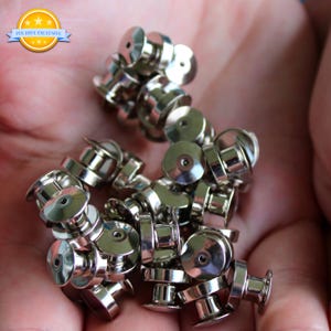 May include: A collection of shiny silver pin backs, displayed against a blurred background. The pin backs have a flat, circular top with a small hole and a post for attaching to pins. The image includes a logo that says "PIN HIVE EXCLUSIVE".