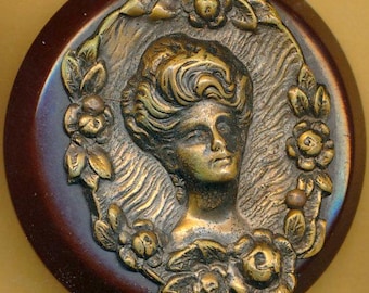May include: A vintage-style button with a dark brown, oval base. The center features a detailed, antique-looking relief of a woman's face surrounded by a floral wreath. The metal relief has a bronze finish, and the button is labeled with the number "#1".