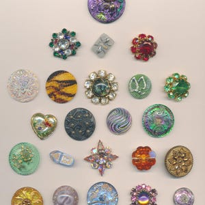 May include: A collection of decorative buttons in various shapes, sizes, and colors. The buttons include floral, geometric, and abstract designs, with embellishments like rhinestones and iridescent finishes. Some buttons are round, oval, or heart-shaped, and feature a range of colors.