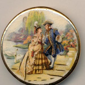 May include: A round, antique porcelain button with a painted scene of a couple in 18th-century attire near a lake. The man wears a blue coat, and the woman is in a yellow and brown dress. The button has a gold-colored rim and a cracked surface.