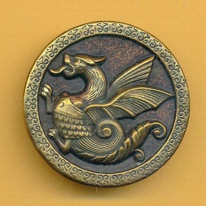 May include: A round brass button featuring a raised relief of a winged dragon with a curled tail. The central figure is set against a dark, textured background, and the outer rim is decorated with a repeating embossed pattern in a metallic gold finish.
