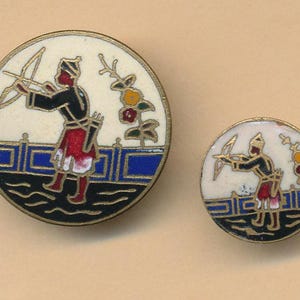 May include: Two round enamel pins. The larger pin features a figure in black and red attire with a bow and arrow, set against a white background with a blue and black border. The smaller pin mirrors the design. Decorative accessories.