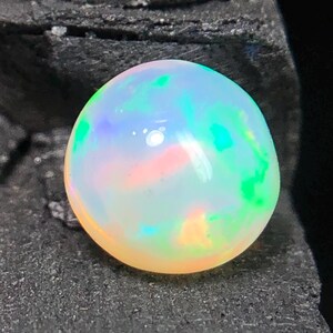 Könnte beinhalten: Ein runder, im Cabochon-Schliff gehaltener Opal-Edelstein mit einer durchscheinenden weißen Basis und lebhaften Farbspielen in Grün, Blau und Orange. Der Stein hat eine glatte, polierte Oberfläche, die das Licht reflektiert und Muster zeigt.