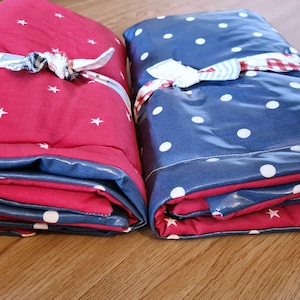 May include: Two folded blankets, one red with white stars, the other blue with white polka dots. Both have a blue border and are tied with a patterned ribbon. The blankets are folded and ready for use.