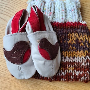 May include: Baby booties with a robin design, in light grey leather with red suede details. The booties are next to a knitted bag with a white handle and a colourful, textured body.