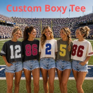 May include: Five boxy t-shirts in black, white, and olive green, each featuring large athletic-style numbers on the front. The shirts have short sleeves and a cropped fit, paired with light blue denim shorts. The text Custom Boxy Tee is visible.
