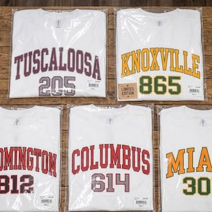 May include: Five white t-shirts in plastic packaging, each with a city name and area code in bold lettering. Cities include Tuscaloosa, Knoxville, Bloomington, Columbus, and Miami. The Knoxville shirt has a "Limited Edition" sticker.