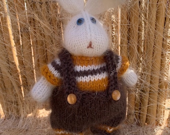 May include: A handmade knitted bunny doll with long white ears and blue eyes. The bunny wears a brown and yellow striped shirt and brown overalls with wooden buttons. The doll is a soft, plush toy.