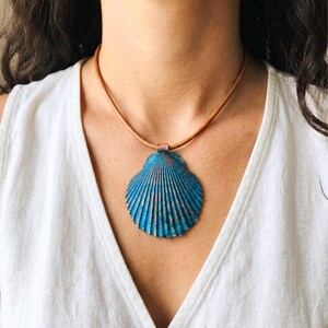 May include: A blue seashell pendant necklace with a brown leather cord. The pendant has a textured surface with a metallic finish. The necklace is worn around the neck.