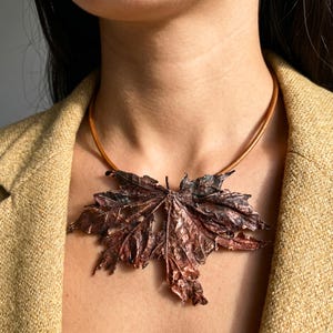 May include: A copper-colored maple leaf pendant necklace. The large, detailed leaf is suspended from a thin, brown leather cord. The necklace is worn against a neutral background, highlighting the intricate details and metallic sheen of the leaf.