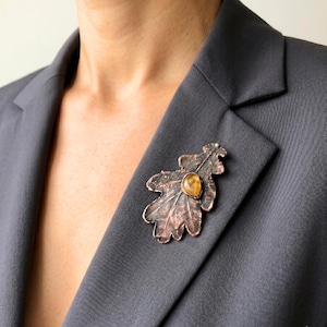May include: A copper-colored leaf-shaped brooch with a central amber-colored stone is pinned to a gray blazer. The brooch has detailed veining and a textured surface, adding a naturalistic element to the garment.