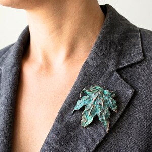 Pode incluir: Broche de folha detalhada em tom de cobre com acabamento esverdeado, presa a um blazer cinza. A broche tem veios intrincados e uma superfície texturizada, criando um design naturalista. A folha tem aproximadamente 7.6 cm de comprimento.