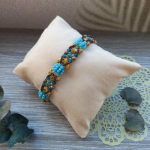 May include: A beaded bracelet featuring a repeating pattern of bright turquoise floral-shaped clusters, interspersed with small metallic bronze beads, dark maroon seed beads, and mottled yellow and brown accent beads in a woven design.
