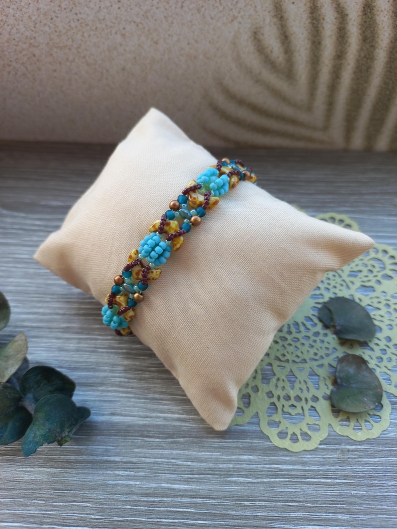 May include: A beaded bracelet featuring a repeating pattern of bright turquoise floral-shaped clusters, interspersed with small metallic bronze beads, dark maroon seed beads, and mottled yellow and brown accent beads in a woven design.