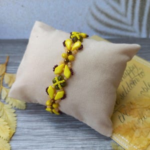Pu&ograve; includere: Un braccialetto di perline con un motivo ripetuto di perline giallo brillante a forma di diamante, piccole perline marrone scuro e perline rotonde verde oliva. Il braccialetto &egrave; realizzato con un design intrecciato che crea un aspetto strutturato.
