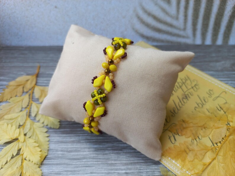 Pu&ograve; includere: Un braccialetto di perline con un motivo ripetuto di perline giallo brillante a forma di diamante, piccole perline marrone scuro e perline rotonde verde oliva. Il braccialetto &egrave; realizzato con un design intrecciato che crea un aspetto strutturato.