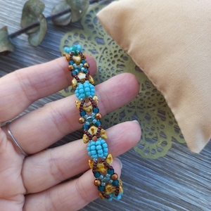 May include: A beaded bracelet featuring a repeating pattern of bright turquoise seed beads, dark brown seed beads, and tan-coloured leaf-shaped beads. The design incorporates metallic bronze accent beads arranged in a structured, woven geometric sequence.