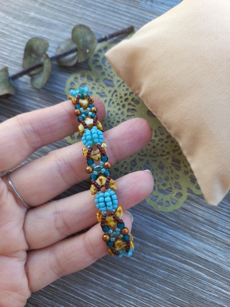 May include: A beaded bracelet featuring a repeating pattern of bright turquoise seed beads, dark brown seed beads, and tan-coloured leaf-shaped beads. The design incorporates metallic bronze accent beads arranged in a structured, woven geometric sequence.