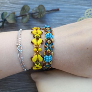 May include: A silver-toned chain bracelet with a small heart charm, alongside two beaded bracelets. One beaded bracelet features yellow diamond-shaped beads and dark brown accents, while the other displays turquoise and brown patterned beads.