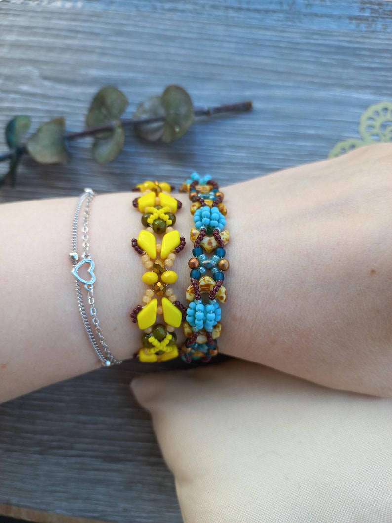 May include: A silver-toned chain bracelet with a small heart charm, alongside two beaded bracelets. One beaded bracelet features yellow diamond-shaped beads and dark brown accents, while the other displays turquoise and brown patterned beads.