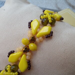 Pu&ograve; includere: Un braccialetto di perline caratterizzato da un motivo ripetitivo di perline giallo brillante a forma di diamante, perline rotonde verde oliva traslucide, piccole perline di conteria marrone scuro e perline rotonde color pesca pallido.