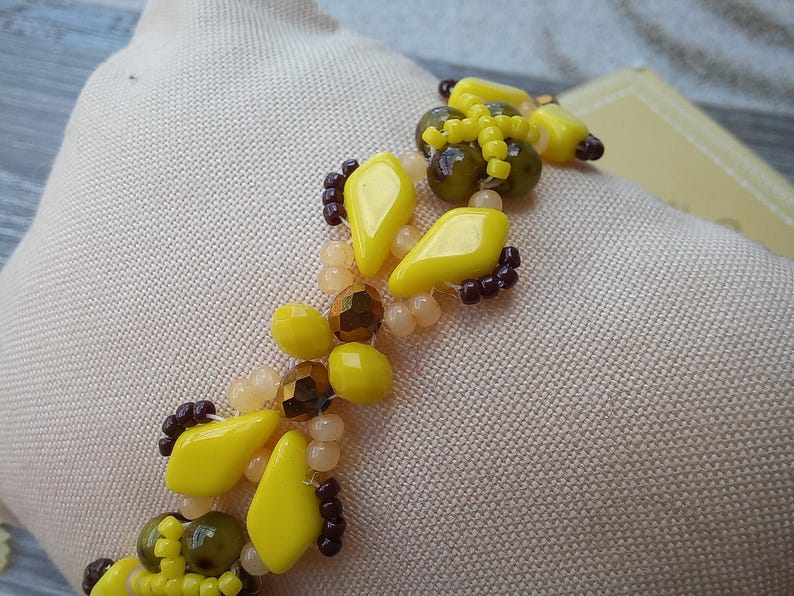 Pu&ograve; includere: Un braccialetto di perline caratterizzato da un motivo ripetitivo di perline giallo brillante a forma di diamante, perline rotonde verde oliva traslucide, piccole perline di conteria marrone scuro e perline rotonde color pesca pallido.