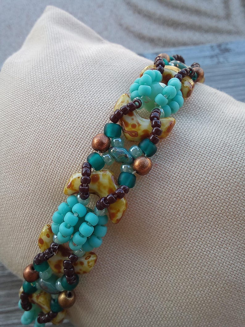 May include: A beaded bracelet featuring a repeating pattern of turquoise blue seed bead clusters, brown seed beads, metallic copper-toned beads, and crescent-shaped beads with a yellow and brown mottled pattern arranged in a woven design.