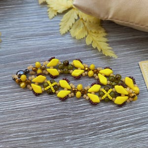 Pu&ograve; includere: Un braccialetto di perline con un motivo ripetuto di perline giallo brillante a forma di diamante, perline rotonde verde oliva traslucide e piccole perline di conteria marrone scuro. Il braccialetto ha un design intrecciato e una chiusura metallica.