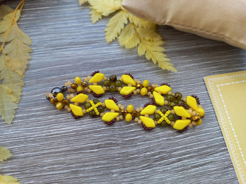 Pu&ograve; includere: Un braccialetto di perline con un motivo ripetuto di perline giallo brillante a forma di diamante, perline rotonde verde oliva traslucide e piccole perline di conteria marrone scuro. Il braccialetto ha un design intrecciato e una chiusura metallica.