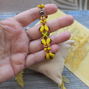 Pu&ograve; includere: Un braccialetto di perline con un motivo ripetuto di perline giallo brillante a forma di diamante, piccole perline arancioni traslucide, perline marrone scuro e una singola perlina sferica grigio metallizzato, intrecciate in un design lineare.