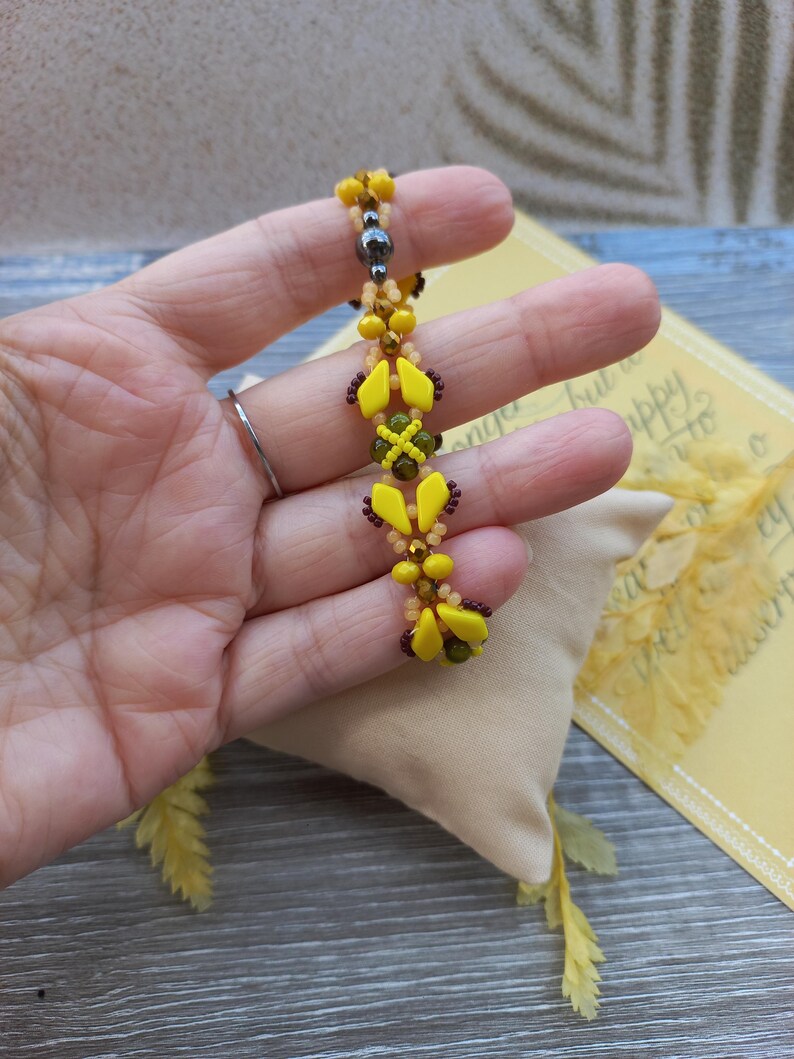 Pu&ograve; includere: Un braccialetto di perline con un motivo ripetuto di perline giallo brillante a forma di diamante, piccole perline arancioni traslucide, perline marrone scuro e una singola perlina sferica grigio metallizzato, intrecciate in un design lineare.