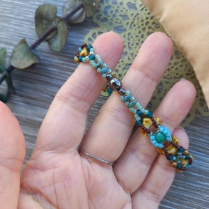May include: A beaded bracelet featuring a repeating pattern of small turquoise, light blue, brown, and yellow beads. The design includes a central dark metallic magnetic clasp, with the beads arranged in a woven, multi-strand structure.