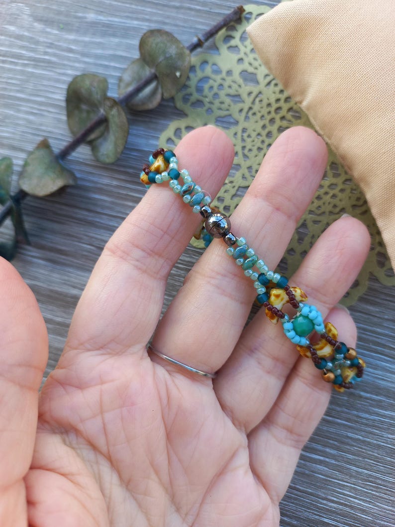 May include: A beaded bracelet featuring a repeating pattern of small turquoise, light blue, brown, and yellow beads. The design includes a central dark metallic magnetic clasp, with the beads arranged in a woven, multi-strand structure.