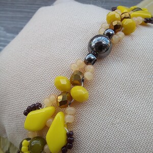 Pu&ograve; includere: Un braccialetto di perline con un mix di perline sfaccettate gialle, verde oliva e bronzo metallizzato, intervallate da piccole perline traslucide color pesca e una singola grande perla sferica liscia grigio metallizzato scuro al centro.