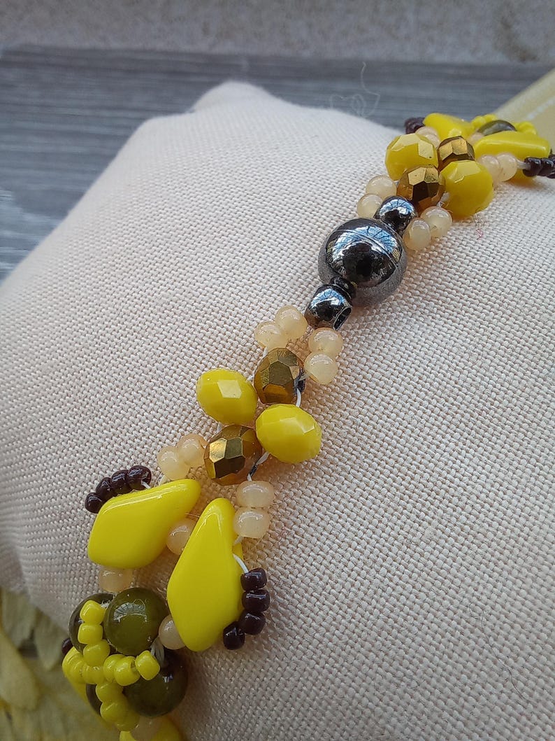 Pu&ograve; includere: Un braccialetto di perline con un mix di perline sfaccettate gialle, verde oliva e bronzo metallizzato, intervallate da piccole perline traslucide color pesca e una singola grande perla sferica liscia grigio metallizzato scuro al centro.