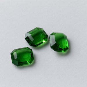 Puede incluir: Tres gemas facetadas con talla esmeralda en un tono verde vibrante. Las gemas se muestran sobre un fondo blanco liso, mostrando su claridad y brillo. Las facetas capturan la luz, creando un efecto brillante.